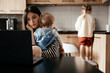 © Ananass - a young mother with many children working on a laptop sitting in the kitchen with a baby in her arms while the eldest daughter helps with cooking and cleaning in the kitchen
