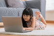 Cute little asian girl uses laptop while prone at the sofa in the living room. Child surfing the internet on computer, browses through internet and watches cartoons online at home