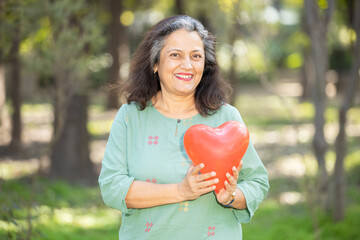 Aging, Portrait of happy Indian Asian senior woman holding red heart balloon at summer park  Elder people heart health care concept. Love, Relationship. 