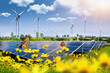 Solar panel with wind turbines against mountains and sky as background and flowers in a blurred foreground.