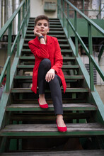 A Stylish Model Girl With Short Brown Hair In A Red Jacket And Pumps, A White T-shirt, Posing For A Photographer Sitting On A Wooden Staircase With Wooden Railings And Propping Her Face With Her Hand