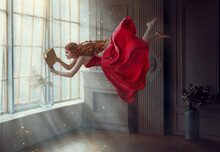 Fantasy Redhead Woman Soars Floats Flies In Air. Art Photo Levitation. Girl Fairy Princess Reads Magic Book, Divine Light From Window. Red Midi Dress, Lon Hair Flutters In Wind. Room Classic Interior