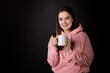 © Alexander - Cute young brunette in a pink hoodie drinking tea or coffee from a large mug, portrait on a black background