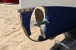 A closeup image of the brown helix of a small fishing boat. The boat is parked in the sand on the beach and is made of wood and is colored white and blue. The boat is a bit old and the wood is worn.