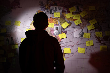 Back view of silhouetted man planning in dark room. Unrecognizable man standing near wall with stickers, photographs and red threads. Planning, conspiracy concept