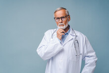 Thoughtful Senior Doctor In Black-rimmed Glasses And White Coat Props Up Chin With Hand. Bearded Man With Stethoscope Stands On Blue Background