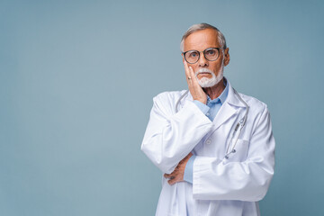 Bearded senior doctor in black-rimmed glasses with stethoscope props up cheek with hand pityingly. Elderly man in white medical coat stands by blue wall