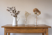 Close Up Of Dried Flowers And Leaves In Glass Bottles And White Jug On Oak Side Table Against Beige Wall (selective Focus)