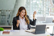 © amnaj - Successful young Asian business woman achieving goals excited raised hands rejoicing with laptop in the office. New startup project concept.