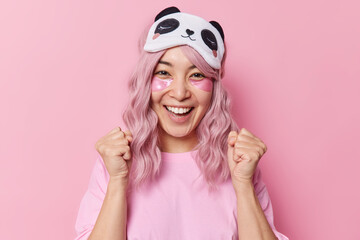 Wall Mural - Horizontal shot of happy pink haired young Asian woman clenches fists celebrates special occasion applies beauty patches under eyes to remove wrinkles wears sleepmask on forehead poses indoor