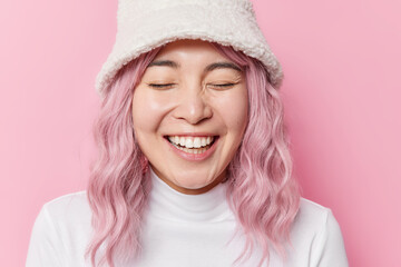 Wall Mural - Cheerful overjoyed Asian woman with rosy hair laughs sincerely shows white teeth keeps eyes closed wears hat and poloneck isolated over pink background. People emotions and feelings concept.