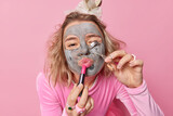 Fototapeta  - Beauty portrait of young female model applies clay mask on face lipstick and curles eyelashes has hairstyle gets ready to party wants to have fabulous fancy look isolated over pink background.