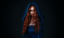 Portrait Fantasy Gothic Red-haired Woman Witch. Vampire Girl In Blue Medieval Dress, Vintage Old Historical Style Hood On Head. Black Background. Red Lips, Hair Flying Soar In Wind. Halloween Costume