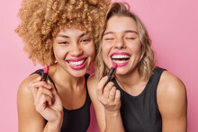 Happy Beautiful Women Apply Lipstick Undergo Beauty Procedures Stand Next To Each Other Dressed In Black T Shirts Isolated Over Pink Background. Two Female Friends Prepare For Party Want To Look Good