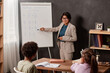 © Mediaphotos - Smiling young Latin teacher pointing at example on whiteboard while explaining new topic to school children at class