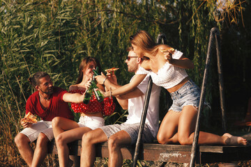 Wall Mural - Group of young people eats pizza and drinking beer on a dock by the river during the summer sunny day