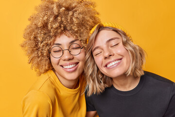 Wall Mural - Portrait of happy young European women keep eyes closed from satisfaction smile toohily being in good mood dressed in casual t shirts express positive emotions isolated over yellow background