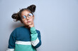 Funny little girl in glasses holding chin doubts, makes decision isolated on yellow studio background