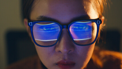 Close up of face of young Asian woman, developer programmer, software engineer, IT support, wearing glasses working hard at night overtime on computer to check coding in bugging system.