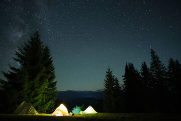 Bright illuminated tourist tents glowing on camping site in dark mountains under night sky with sparkling stars. Active lifestyle concept