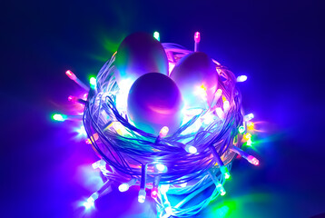 Creative conceptual photo design of 3 Easter eggs in bird nest made with glowing colorful decoration led light bulbs and electric wire on dark black background and copy space. Close up macro top view.