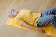 © MyJuly - a man is lying on the floor in the shavasana pose and meditating. does yoga and fitness in the gym on a yellow mat. senior yoga at home or in the gym for training. a fifty-year-old man with gray hair