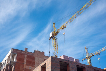 Two high-rise construction cranes and unfinished monolithic building on the blue sky background. Building construction site with cranes.