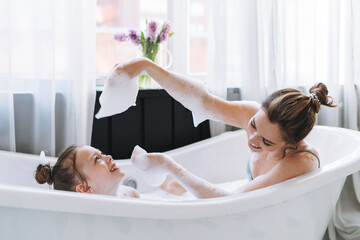 Young mother woman with long hair with little tween girl daughter in pajamas having fun in the bath with foam at home