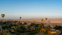 Bright Balloons Over Luxor. Village Houses, Palm Trees, Green Vegetation Are Visible Below. In The Distance, In The Sandy Desert - Landing Balloons, Cars. Blue Sky. Egypt