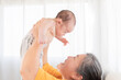 © Rakchanok - Side view grandmother and newborn baby play together at home, Grandma holding adorable infant in air and looking with love and tender. Spend time together. Senior women making funny faces to toddler.