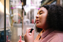 Young Woman With Curly Hair Applying Lip Gloss In Front Of Store Window