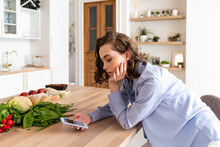 Woman Using Mobile Phone By Vegetables At Dining Table In Kitchen