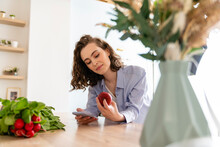 Young Woman Checking Calories Of Apple On Mobile Phone In Kitchen
