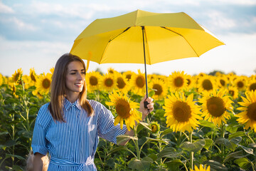 Young woman in a dress hides flowers from the sun under a yellow umbrella on a sunflower field