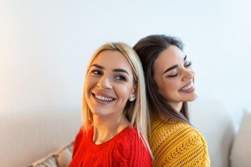 Two beautiful women with blond and brunette hair wearing knitted jumpers sitting on the sofa and smilling, Cheerful women back against back looking at camera and sitting on the sofa