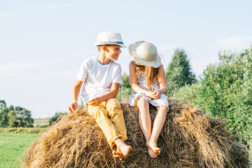 Portrait of two barefoot children boy and girl sitting on haystack in field. Light sunny day. First love concept. Smiling and laughing best friends wearing hats. Trees on background.