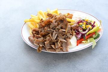 Thin slices of doner kebab meat with french fries, salad and yogurt dip sauce on a plate and a gray table, copy space, selected focus