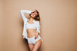 © F8  \ Suport Ukraine - Attractive woman with slender figure and natural tan in white cotton underwear touches leg on solid beige color text space background. Skin care concept