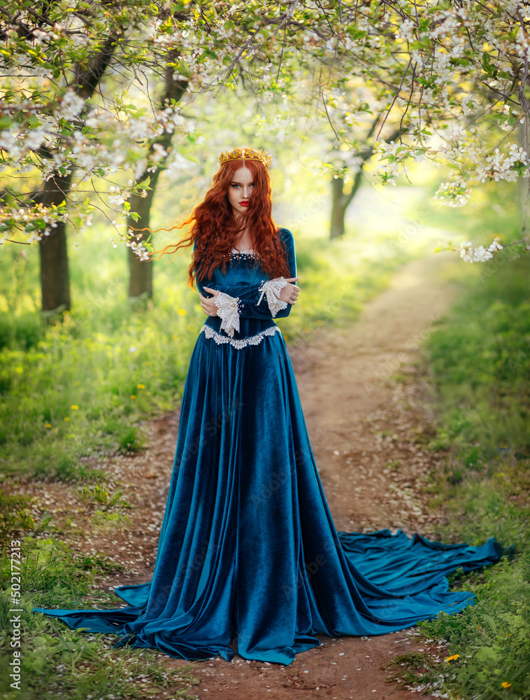 Photo Stock Redhead fantasy woman queen. Blue long velvet medieval dress,  vintage clothing. Red curly hair flying waving in wind. Summer nature green  flowering trees garden footpath path. Girl Princess walking |