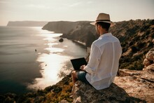 Digital Nomad, Man In The Hat, A Businessman With A Laptop Sits On The Rocks By The Sea During Sunset, Makes A Business Transaction Online From A Distance. Remote Work On Vacation.