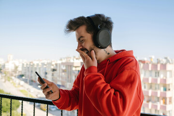 Wall Mural - Laughing, the young man laughs loudly at the message he receives, young man wearing headphones on balcony with phone in hand, funny moment and smile. face expression