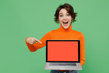 Young smiling happy woman 20s wear orange turtleneck hold use work point finger on laptop pc computer with blank screen workspace area isolated on plain pastel light green background studio portrait