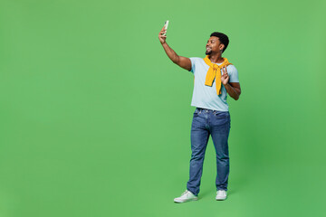 Wall Mural - Full body young man of African American ethnicity wear blue t-shirt doing selfie shot on mobile cell phone post photo on social network show v-sign isolated on plain green background studio portrait