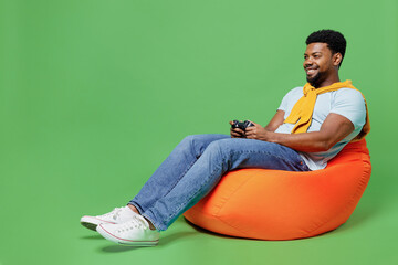 Wall Mural - Full body smiling fun young man of African American ethnicity 20s wear blue t-shirt sit in bag chair hold in hand play pc game with joystick console isolated on plain green background studio portrait