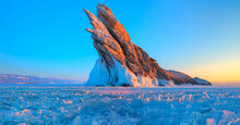 A Granite Rock With Steep Slopes Rises Above A Frozen Lake With Reflection On The Ice At Sunset -  Lake Baikal, Russia