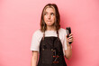 Young hairdresser woman holding scissors isolated on pink background confused, feels doubtful and unsure.