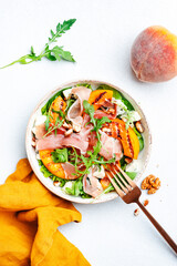 Wall Mural - Summer salad bowl with sweet grilled peach, jamon, soft cheese, walnuts and fresh arugula on white kitchen table background, top view, negative space