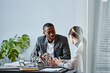 © Seventyfour - Portrait of young black businessman talking to female colleague during meeting in office against pale blue wall, copy space