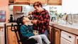 Latin mother taking care of son with disability on wheelchair inside home kitchen - Focus on mom face