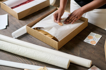 Hands of young black woman putting sticker on top of wrapping paper while packing folded clothes into cardboard box by table